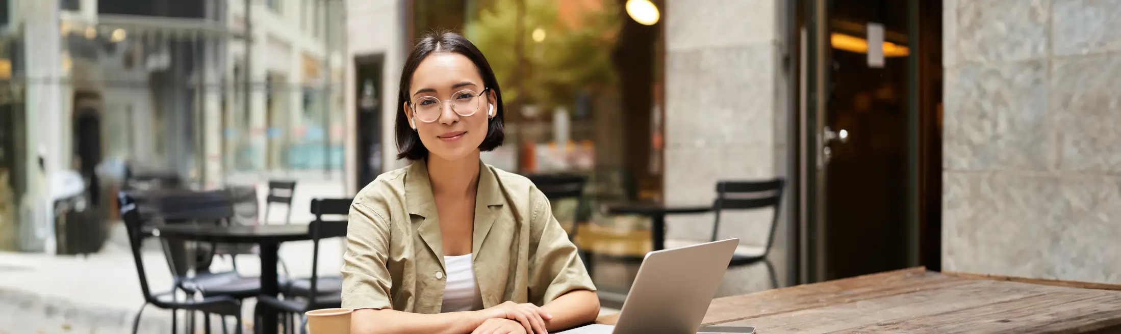 young-woman-student-working-from-cafe-sitting-with-laptop-cup-coffee-outdoors-looking